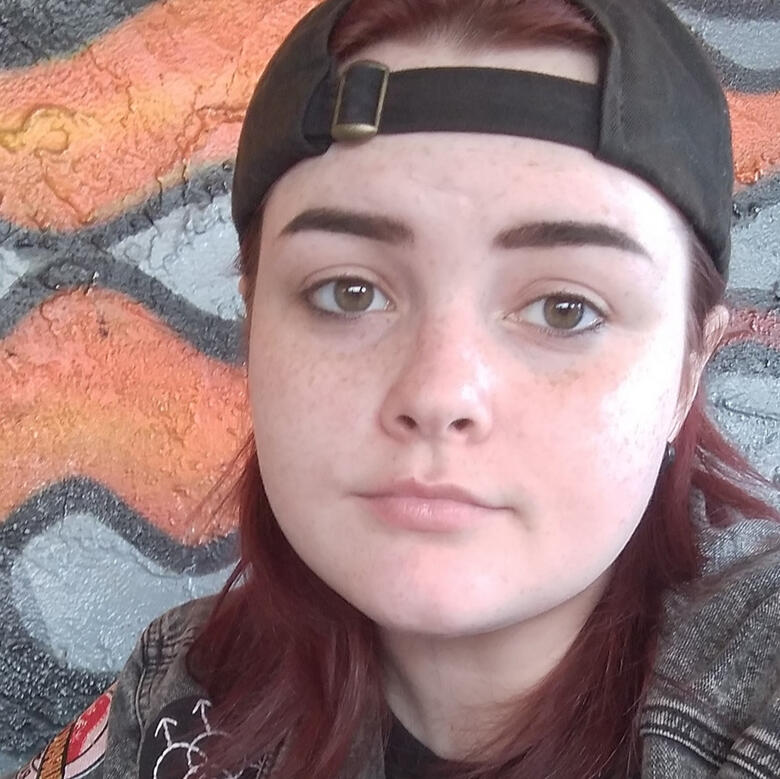 a selfie of me in front of a flame mural wall, i have on a black cap with burgundy red hair, green eyes and black smudged liner on my bottom lids, my eyebrows are groomed and im wearing a black shirt and denim jacket covered in homemade patches